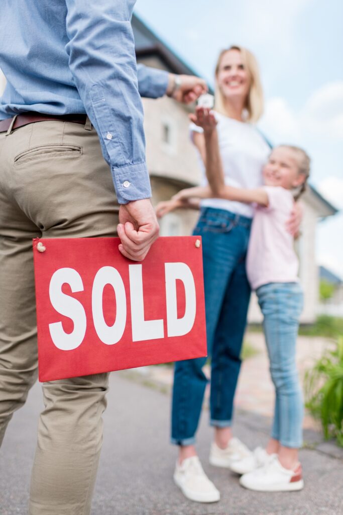 cropped image of realtor with sold sign giving key to young woman with daughter in front of new